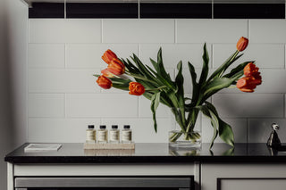 A modern kitchen countertop with a vase of vibrant orange tulips and a set of fragrance oils Australia displayed neatly, against a sleek white tiled backsplash.